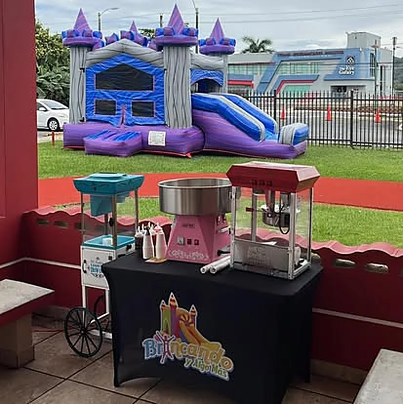 Mesa con el mantel de Brincando y Algo Más y las tres máquinas montadas: algodón de azúcar, piragua y pop corn, con un castillo inflable con chorrera armado al fondo
